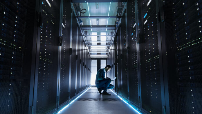 IT Technician Works on Laptop in Big Data Center full of Rack Servers. He Runs Diagnostics and Maintenance.